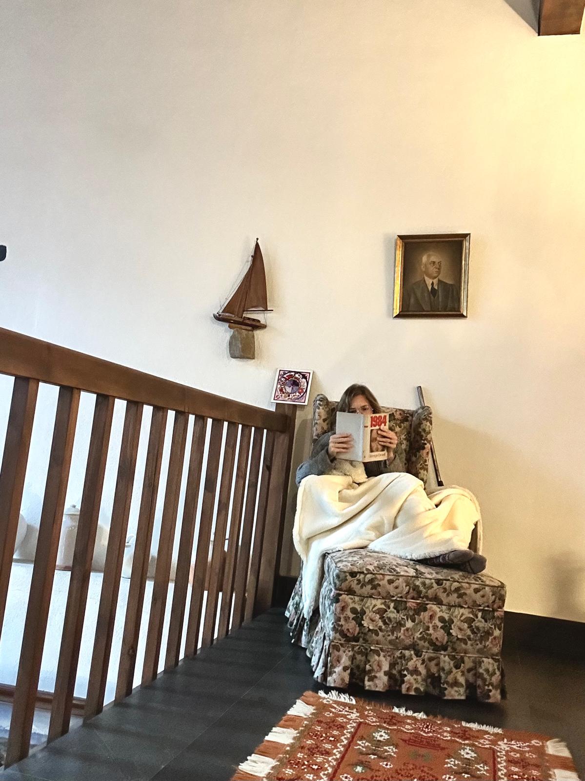 “Ana leyendo junto a la barandilla de madera en Casa Goikomaia, una escena cálida y lenta que invita a parar.”